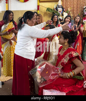 In einem hinduistischen Tempel eine ältere Frau besprüht Blütenblätter auf den Köpfen einer Braut & Bräutigam bei einer Trauung. In Queens, New York City. Stockfoto