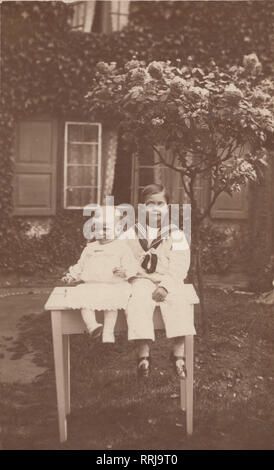 Jahrgang fotografische Postkarte zeigt zwei Kinder Satat auf einen Tisch im Garten. Der ältere Junge trägt einen Matrosen Outfit. Stockfoto