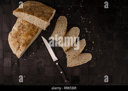 Hausgemachtes Brot, einfache und gesunde Ernährung. Das Brot mit verschiedenen Scheiben und einem Messer geschnitten, von oben auf einem schwarzen Hintergrund gesehen Stockfoto