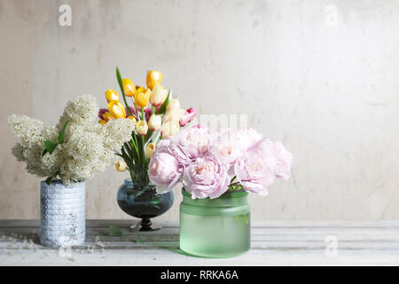 Anordnung der Blumen, lila Pfingstrosen, Tulpen, steht auf alte hölzerne Bretter gegen eine Wand, in Gläser in verschiedenen Farben Stockfoto