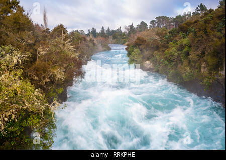 Huka Falls auf dem Waikata Fluss, Wairakei National Park, Neuseeland. Reißenden Strom fließt durch die von Bäumen gesäumten Schlucht mit bewölktem Himmel Stockfoto