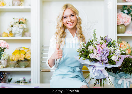 Schöne lächelnd weibliche Flower Shop Besitzer sittng auf Zähler und Holding Bouquet und zeigt mit dem Daumen nach oben anmelden Stockfoto