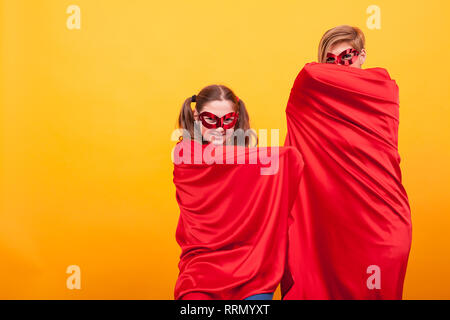 Schöne Mutter mit ihrer Tochter wie Superheldinnen versteckt unter ihren roten Umhang über der Hintergrund gelb gekleidet. Rote Maske. Gute Eltern. Stockfoto