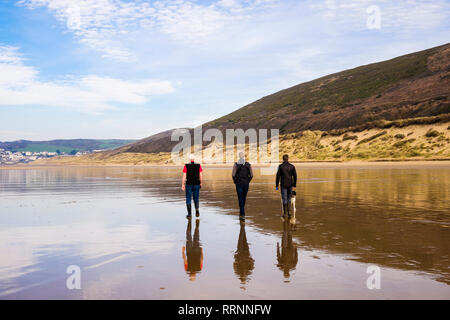 Drei Männer mit einem Hund sich im nassen Sand zu Fuß an einem ruhigen Strand bei Ebbe. Woolacombe, North Devon, England, Großbritannien, Großbritannien Stockfoto