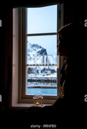 Nachdenkliche Frau, die auf der Suche Fenster bei Snowy Mountain, Reine, Lofoten, Norwegen Stockfoto