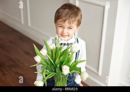 Süße Junge in ein weißes Hemd und eine Quaste Schmetterling gibt einen Blumenstrauß aus den weißen Tulpen Stockfoto