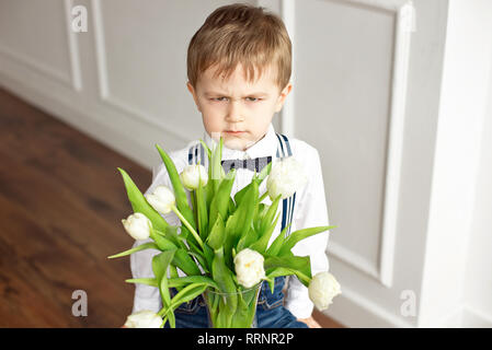 Süße Junge in ein weißes Hemd und eine Quaste Schmetterling gibt einen Blumenstrauß aus den weißen Tulpen Stockfoto