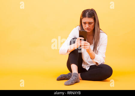 Mädchen suchen auf Ihr Telefon besorgt während Sie auf gelben Hintergrund sitzen. Gesichtsausdruck Stockfoto