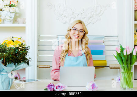 Schöne weibliche Blüte shop besitzer in Gläsern am Schreibtisch sitzen, mit Blick auf die Kamera und Laptop Stockfoto