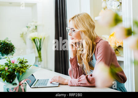 Schöne nachdenkliche weiblichen Blumenhändler in Gläser mit Laptop in Flower Shop Stockfoto