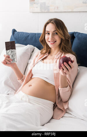 Lächelnd schwangere Frau mit Schokolade und Apple im Bett Stockfoto