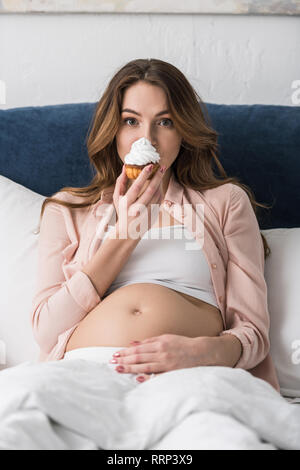 Attraktive schwangere Frau im Bett liegen und essen Kuchen Stockfoto