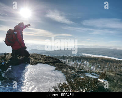 Stehende Mann mit angehobenem Arm auf eisigen Stein und Blick auf schneebedeckte Berge. Landschaft mit Reisende, Stockfoto