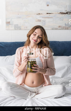 Lächelnd schwangere Frau essen Pickles auf dem Bett Stockfoto