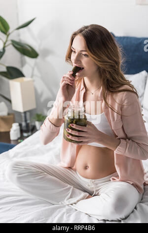 Brunette schwangere Frau im Bett essen Pickles Stockfoto