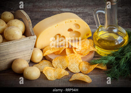 Knusprige Kartoffelchips mit Käse, Kartoffeln in Holzkiste und Öl zum Braten am Küchentisch. Stockfoto