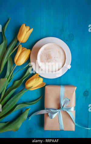 Kaffeebecher mit gelben Tulpen Blumen und Geschenk Box auf Blau rustikal Tabelle von oben, Frühstück am Muttertag oder Tag der Frauen Stockfoto