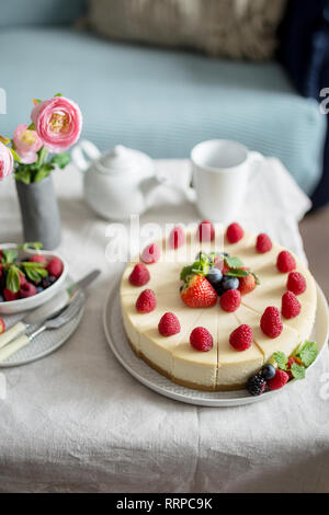Käsekuchen, hausgemachten Kuchen mit Erdbeeren Beeren in eine graue Platte, mit Schale, Teekanne, Gabel, Messer, frische Beeren, Vase mit Blumen, Seitenansicht. Süßigkeiten Stockfoto