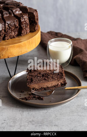 Stück Schokoladenkuchen auf graue Platte mit Gabel, Glas Milch, Servietten, stand mit Schokoladenkuchen auf grauem Hintergrund. Konzept der gesunden Ernährung. Vertica Stockfoto