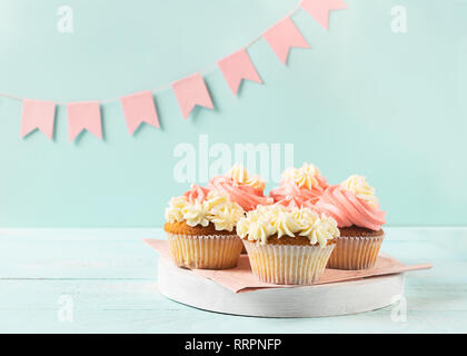 Muffins sind mit dicken weißen und rosa Creme auf einem hellblauen Hintergrund rosa festliches Flaggen dekoriert. Festliche Konzept. Stockfoto