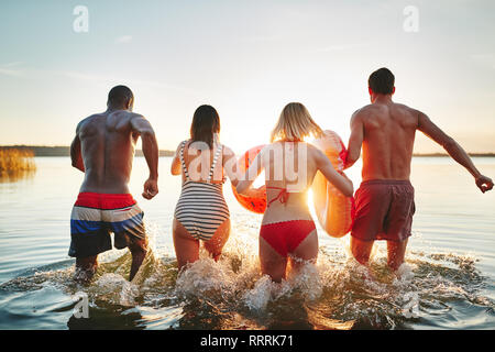 Die aussenspiegel von zwei jungen Paaren in Badebekleidung Spritzwasser während in einen See zusammen laufen bei Sonnenuntergang Stockfoto