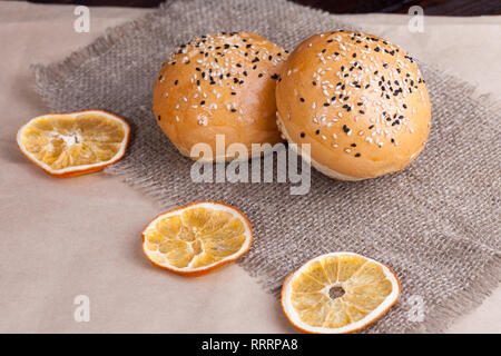 Heiße süße Brötchen auf einem urigen Hintergrund. Geformte Essen mit Sesam auf Gewebe und trockene Apfelsinen. Stockfoto