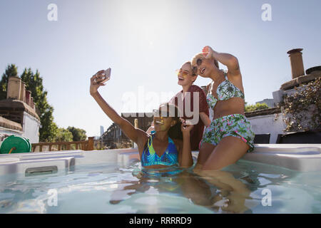 Glückliche, unbeschwerte junge Frauen Freunde in Bikinis unter selfie mit Kamera Handy in sonniger, Whirlpool auf dem Dach Stockfoto