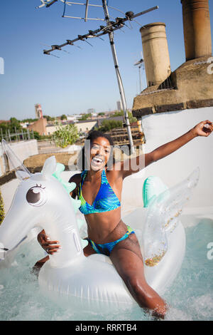 Portrait verspielt, unbeschwerte junge Frau im Bikini auf aufblasbaren Pegasus im sonnigen Dachterrasse mit Whirlpool Stockfoto