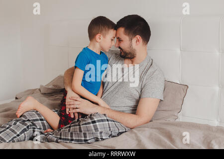 Portrait von mittleren Alter kaukasischen Vater im Gespräch mit jungen Sohn. Mann übergeordneten Holding umarmen mit Kind auf Bett im Schlafzimmer zu Hause. Authentischen Lebensstil Stockfoto