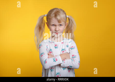 Cute grumpy wütend Kaukasischen blonde Mädchen mit blauen Augen in weißem Kleid posiert im Studio auf gelbem Hintergrund mit Arme auf der Brust verschränkt. Kid Kind Stockfoto