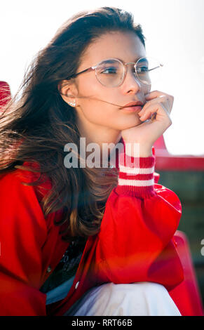 Jungen tennager Girl Modell in den Sitz eines Baseball Stadium das Tragen der roten Bomber Jacke, hipster gerundete Gläser, weisse Hosen und Turnschuhe Stockfoto