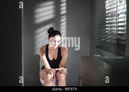 Porträt einer lächelnde Frau mit negativer Schwangerschaftstest Kit, auf der Toilette, zu Hause zu sitzen. Gerne jungen Latino Frau immer gute Nachrichten. Stockfoto