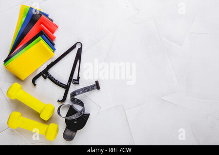 Ansicht von oben Bild von Kurzhanteln, bunte elastische Gummi Expander, Bremssättel und Maßband auf einem weißen strukturierten Hintergrund. Gesunder Lebensstil, Fitnessraum und nicht Stockfoto