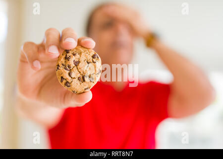 Mittleres Alter Mann essen Chocolate Chips cookies zu Hause mit der Hand betonte am Kopf, mit Scham und Überraschung Gesicht schockiert, wütend und frustriert. Angst und Stockfoto