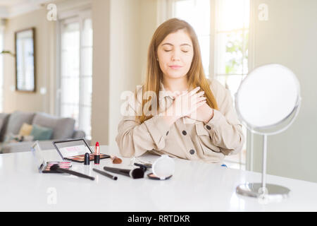 Schöne junge Frau mit Make up Kosmetika lächelnd mit der Hand auf der Brust mit geschlossenen Augen und dankbare Geste auf das Gesicht. Gesundheit Konzept. Stockfoto