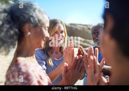 Freunde mit gefalteten Händen in Kreis an sonnigen Strand während Yoga Retreat Stockfoto