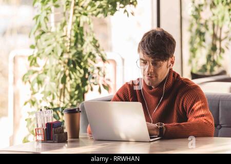 Lächelnd Mann hören Musik im Kopfhörer und mit Laptop in der Nähe holding Einweg Becher in Cafe Stockfoto