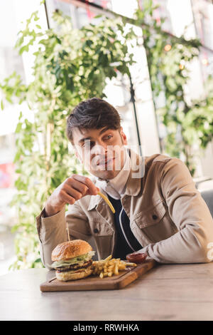 Hübscher junger Mann mit Pommes frites in der Nähe lecker Burger auf Schneidebrett im Cafe Stockfoto