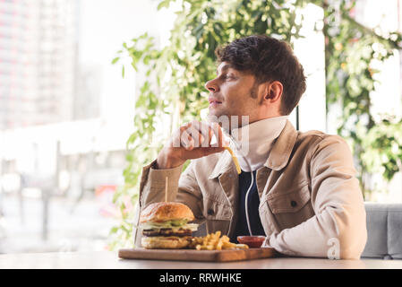 Fröhlicher junger Mann mit Pommes frites in der Nähe lecker Burger auf Schneidebrett im Cafe Stockfoto