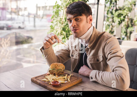 Freundliche Mann mit Pommes frites in der Nähe von köstlichen Burger auf Schneidebrett im Cafe Stockfoto