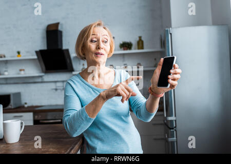 Überrascht ältere Frau mit Finger auf Smartphone zu Hause zeigen Stockfoto