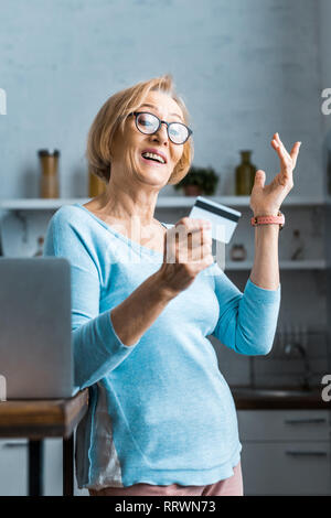 Überrascht ältere Frau in Gläsern und deutete mit der Hand und hält Kreditkarte in der Nähe von Laptop zu Hause Stockfoto