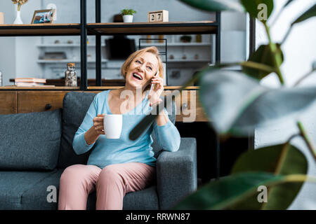 Lachen ältere Frau mit Tasse Kaffee sitzen auf der Couch und Reden auf dem Smartphone im Wohnzimmer Stockfoto