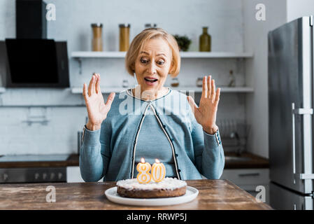 Aufgeregt senior Frau sitzt und Kuchen mit '80' Zeichen auf während der Geburtstagsfeier Stockfoto