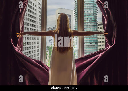 Junge Frau öffnet das Fenster Vorhänge und schaut auf die Wolkenkratzer in der großen Stadt Stockfoto