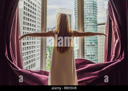 Junge Frau öffnet das Fenster Vorhänge und schaut auf die Wolkenkratzer in der großen Stadt Stockfoto