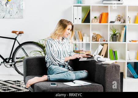 Pretty Woman im gestreiften Hemd sitzt auf einem Sofa und mit Laptop Stockfoto