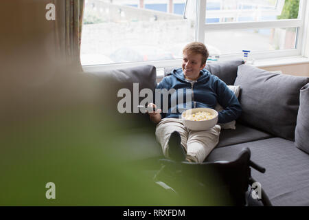 Junge Frau, Fernsehen und essen Popcorn auf dem Sofa mit Füßen auf Rollstuhl Stockfoto