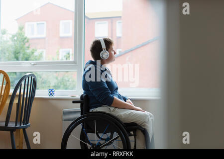 Nachdenkliche junge Frau im Rollstuhl Musikhören mit Kopfhörern mit Fenster Stockfoto