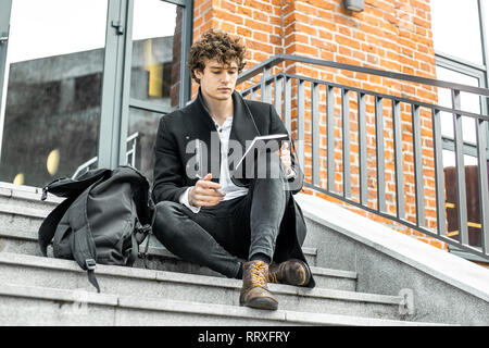 Attraktiver Mann im schwarzen Mantel sitzen auf konkrete Schritte der modernen roten gemauerte Gebäude und Schreiben Notizen in seinem Notizblock. Stockfoto